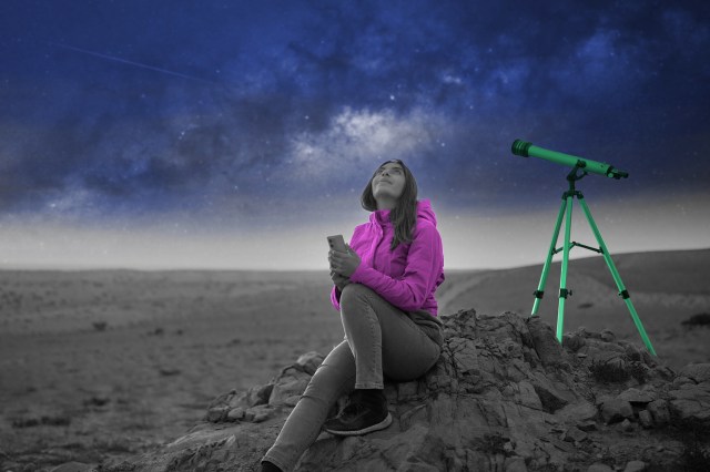 Woman holding phone gazing up at night sky beside telescope